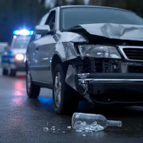 Accidents Caused by Drunk Drivers Representation A damaged car with a broken alcohol bottle on the ground and police lights in the background, representing the impact of drunk driving accidents and legal accountability.
